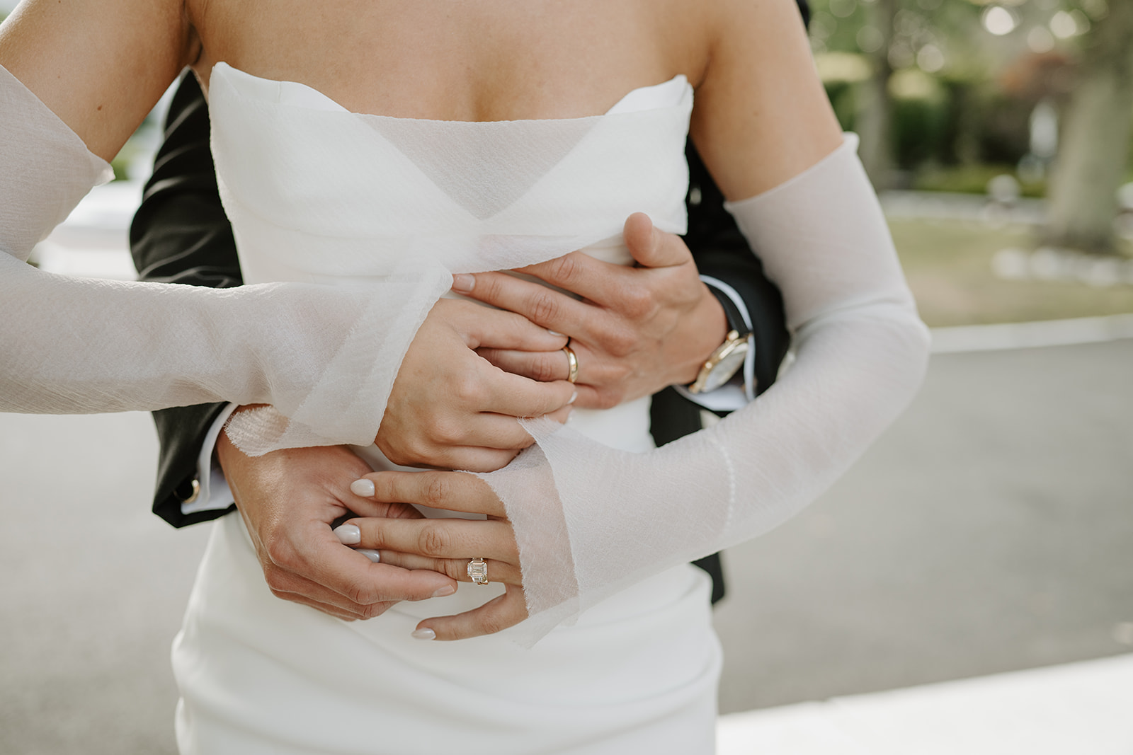 Close-up of the groom holding the bride from behind, showcasing their wedding rings and sheer gloves.